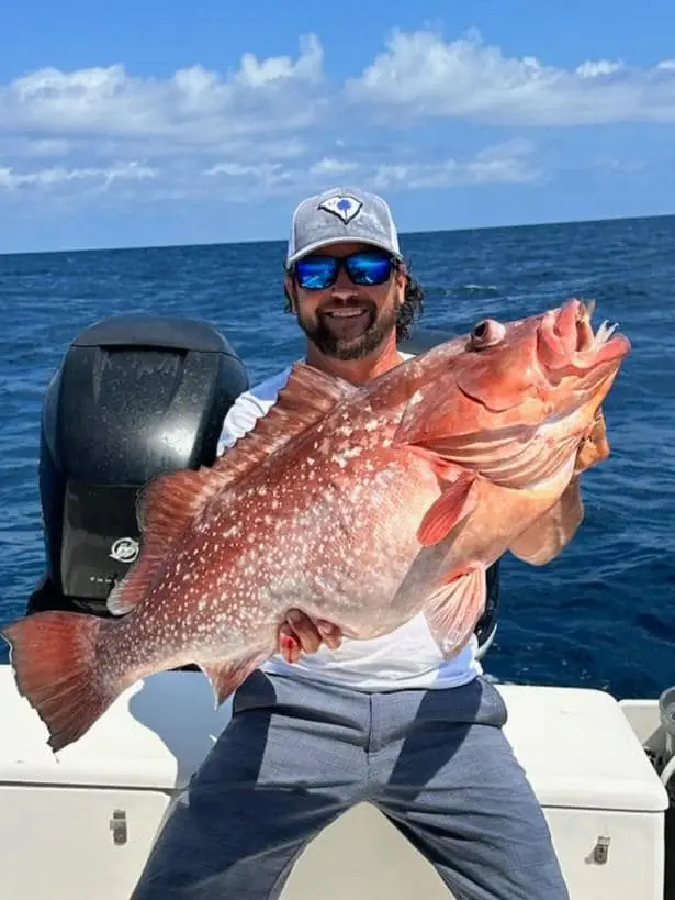 Man proudly holds a large red grouper fish while fishing at sea.